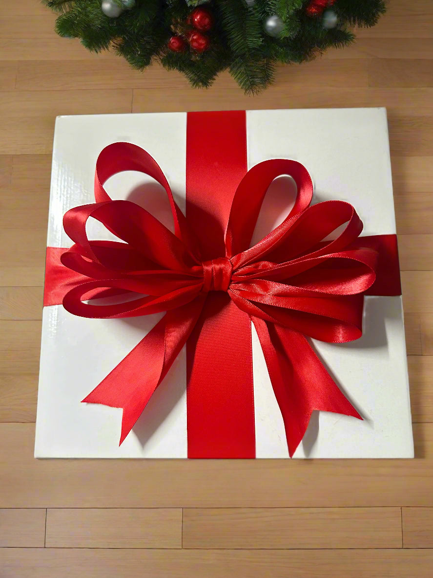 White gift box with a large red bow on a wooden floor, with a Christmas wreath in the background.